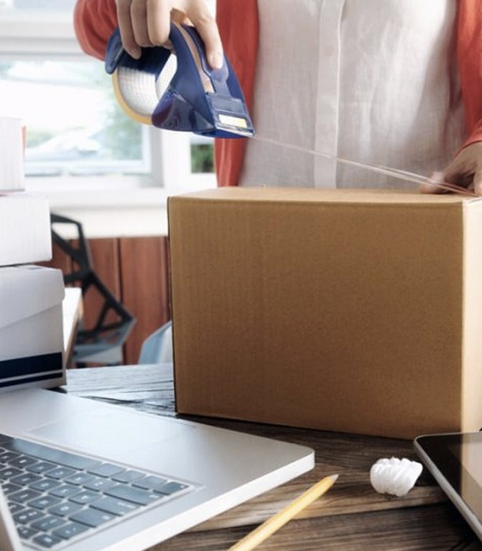 Person sealing a cardboard parcel with packing tape beside a laptop