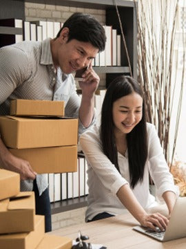Two people preparing online orders beside stacked parcels and a laptop